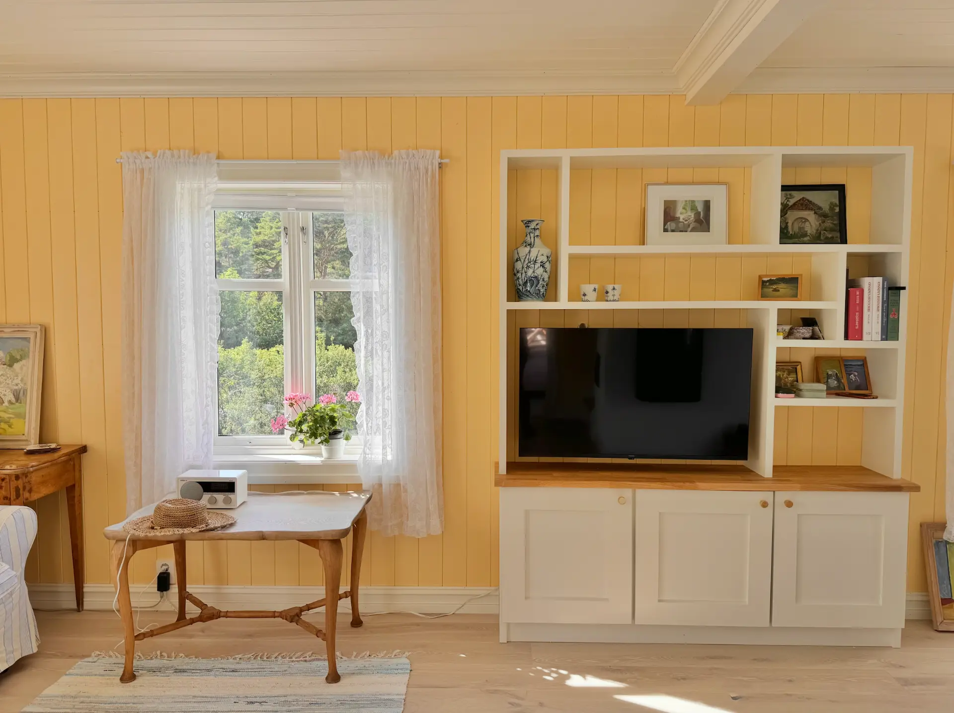 Stue med gul panelvegg, blondegardiner og innebygd reol